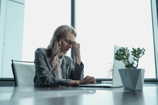 A picture of a woman at an office with a laptop, experiencing a headache and holding her head
